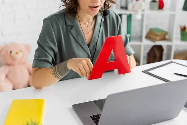 Ausgeschnittene Ansicht eines tätowierten Logopäden im Headset, der während des Online-Unterrichts am Laptop spricht und Buchstaben a hält — Stockfoto