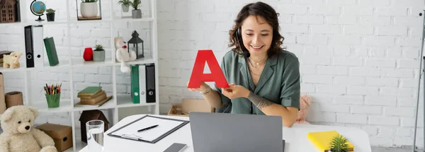 Positive speech therapist in headset holding letter a during video lesson on laptop in consulting room, banner — Stock Photo