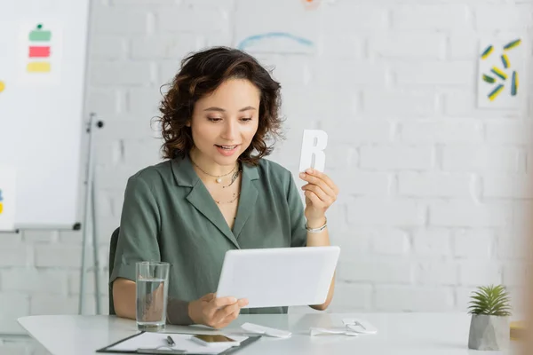 Speech therapist holding letter during video call on digital tablet near smartphone and clipboard in consulting room — Stock Photo