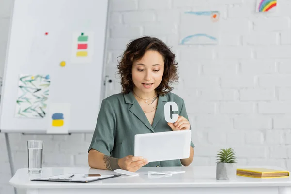 Tattooed speech therapist holding letter c during lesson on digital tablet in consulting room — Stock Photo