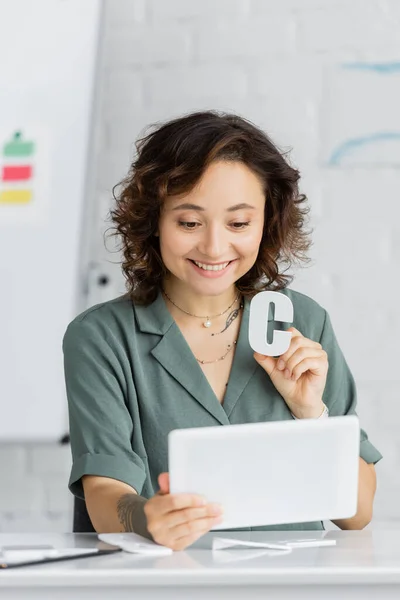Terapeuta del habla positiva sosteniendo carta y tableta digital durante la lección en línea en la sala de consulta - foto de stock