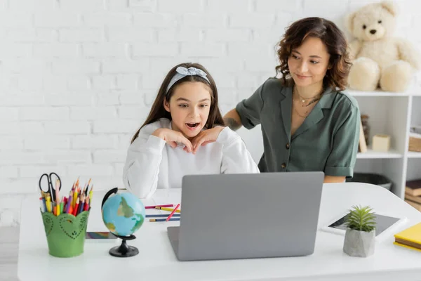 Девочка-подросток смотрит на ноутбук во время онлайн-урока логопедии рядом с мамой дома — стоковое фото