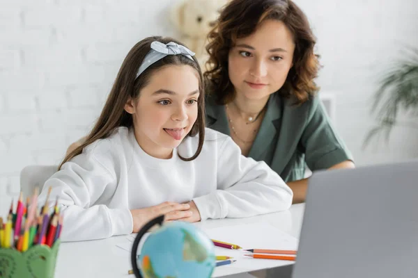 Preteen girl sticking out tongue during speech therapy online lesson near mom at home — Stock Photo