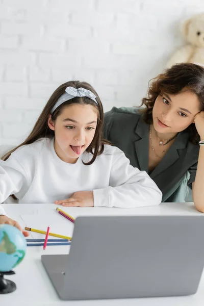 Ребенок высовывает язык во время урока логопедии онлайн рядом с родителем дома — стоковое фото