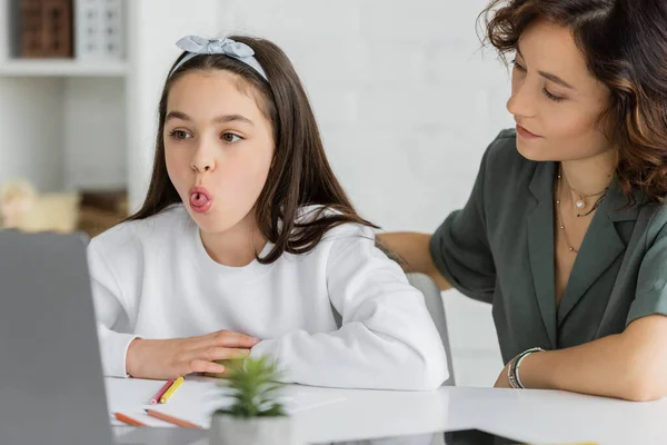 Жінка, дивлячись на дочку, що стирчить язиком під час уроку мовної терапії на ноутбуці вдома — стокове фото
