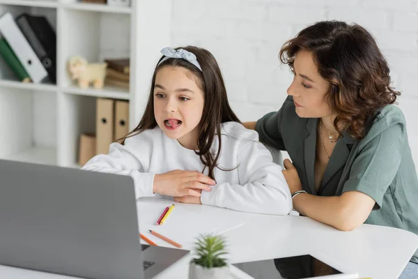Parent sitting near daughter having speech therapy online lesson near devices at home — Stock Photo