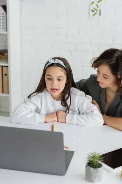 Улыбающаяся мама смотрит, как дочь разговаривает во время урока логопедии на ноутбуке дома — стоковое фото