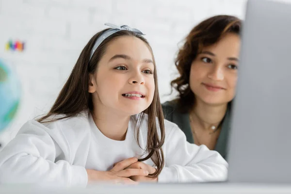 Улыбающийся ребенок с логопедией онлайн урок на ноутбуке рядом с размытым родителем дома — стоковое фото