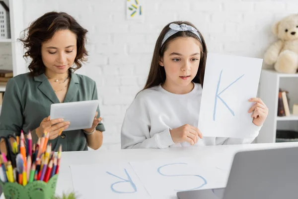Preteen menina segurando papel com letra k durante a terapia da fala aula on-line no laptop perto da mãe em casa — Fotografia de Stock
