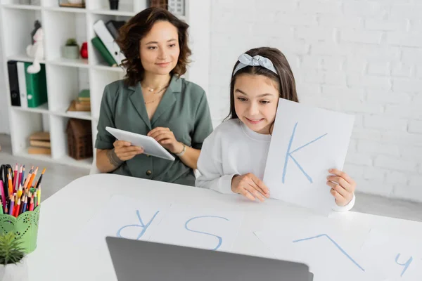Sonriente chica sosteniendo papel con carta durante la lección de terapia del habla en el ordenador portátil cerca de mamá con tableta digital en casa - foto de stock