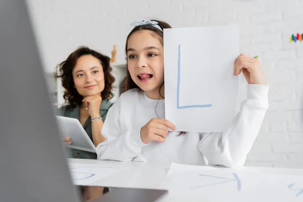 Ребенок высовывает язык и держит бумагу с письмом во время урока логопедии на ноутбуке рядом с мамой дома — стоковое фото
