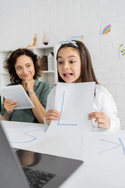Menina segurando papel com carta durante a terapia da fala vídeo lição perto de sorrir mãe com tablet digital em casa — Fotografia de Stock