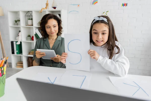 Niño preadolescente sonriendo y sosteniendo papel con carta durante la lección de terapia del habla en la computadora portátil cerca de los padres en casa - foto de stock