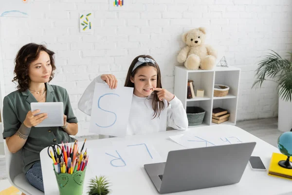 Sonriente niño sosteniendo papel con letras s y apuntando a la boca durante la terapia del habla lección en línea cerca de mamá en casa - foto de stock