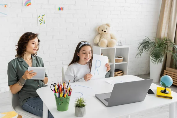Menina positiva segurando papel com carta durante a aula de terapia da fala no laptop perto da mãe com tablet digital em casa — Fotografia de Stock