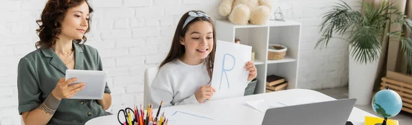 Улыбаясь ребенок-подросток держит бумагу с письмом во время урока логопедии видео на ноутбуке рядом с мамой дома, баннер — стоковое фото