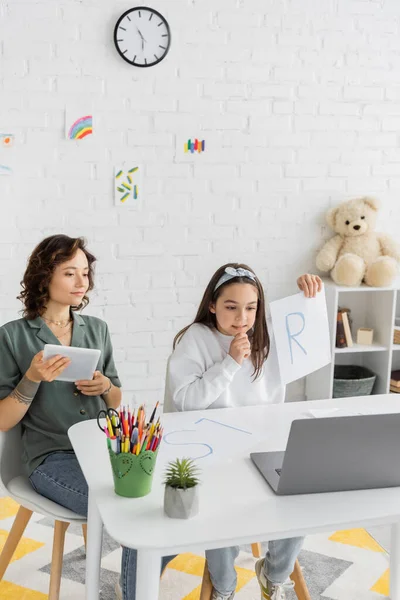 Papel positivo para criança segurando com carta e conversando durante a aula de terapia da fala on-line perto da mãe em casa — Fotografia de Stock