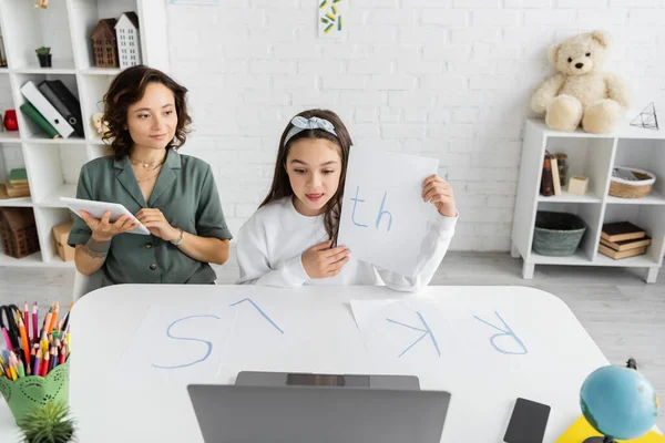 Улыбающаяся женщина с помощью цифрового планшета рядом с дочерью проведение бумаги с буквами й во время логопедии онлайн урок дома — стоковое фото