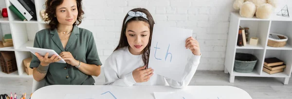 Preteen menina segurando papel com letras perto de sorrir mãe usando tablet digital em casa, banner — Fotografia de Stock