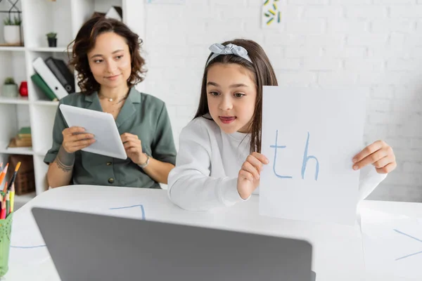 Preteen child holding paper with letters near laptop and mom during online speech therapy at home — Stock Photo
