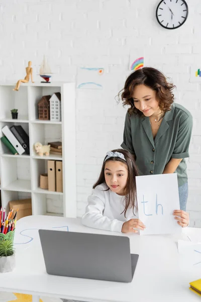 Sonriente madre de pie cerca de la hija sosteniendo papel con cartas durante la terapia del habla en línea lección en casa - foto de stock