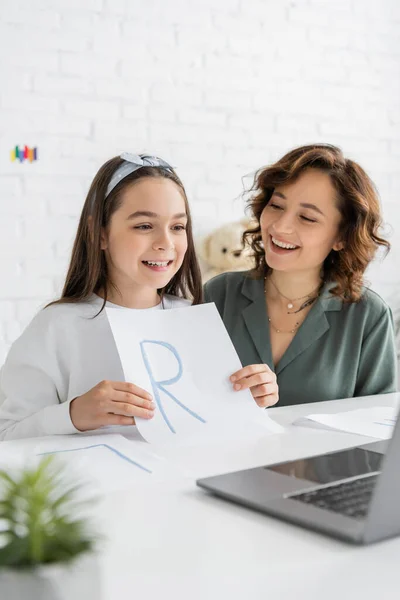 Happy kid holding paper with letter during speech therapy video lesson near parent at home — Stock Photo