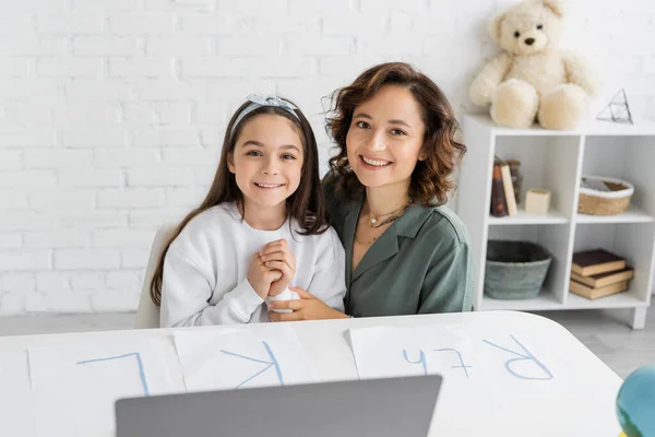 Cheerful woman and daughter looking at camera near papers with letters on desk — Stock Photo