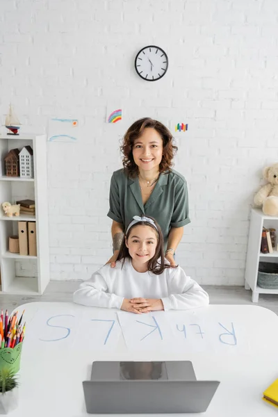 Smiling mother and preteen daughter looking at camera near laptop papers with letters on desk — Stock Photo