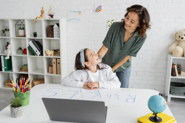 Smiling kid looking at mom near papers with letters and laptop at home — Stock Photo