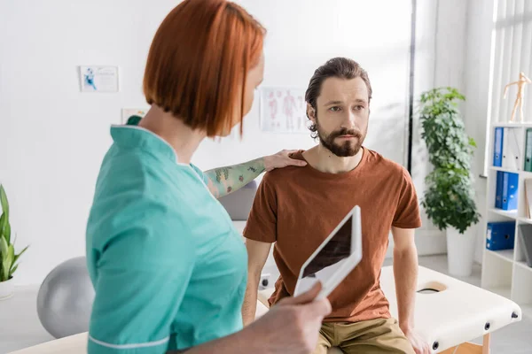 Redhead physiotherapist with digital tablet calming upset man during appointment in rehabilitation center — Stock Photo