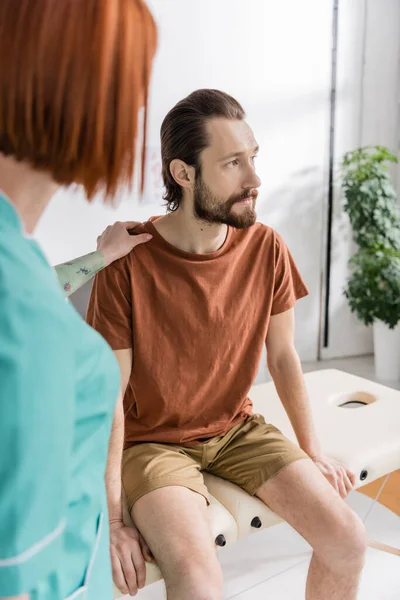 Розмитий фізіотерапевт заспокоює незадоволеного бородатого чоловіка, який сидить на масажному столі в консалтинговій кімнаті — стокове фото