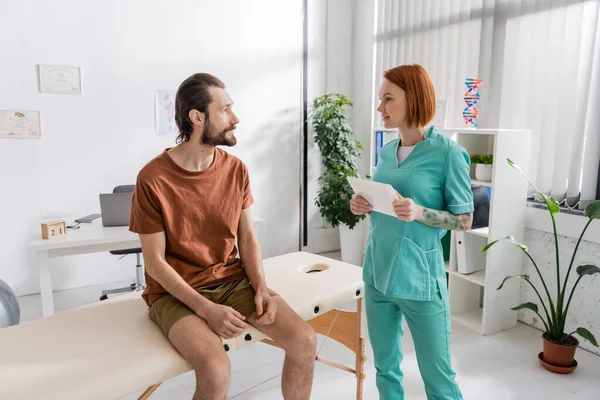 Redhead rehabilitation specialist with digital tablet talking to bearded man sitting on massage table in consulting room — Stock Photo