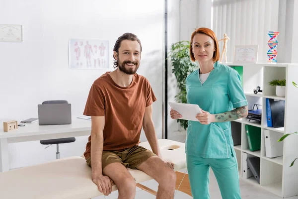 Happy bearded man and physiotherapist with digital tablet smiling at camera in rehabilitation center — Stock Photo