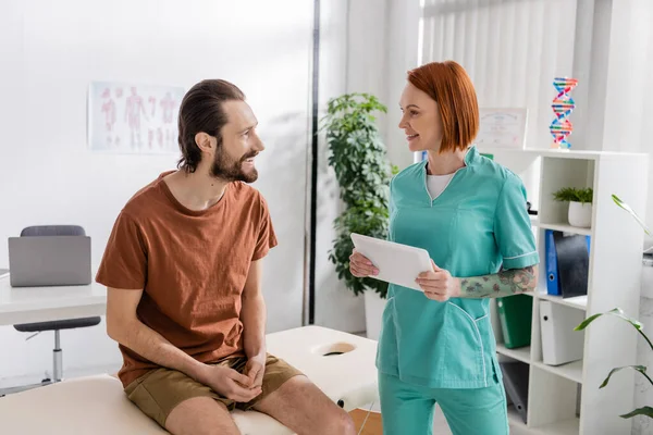 Positive bearded man sitting on massage table and talking to redhead physiotherapist with digital tablet in rehab center — Stock Photo