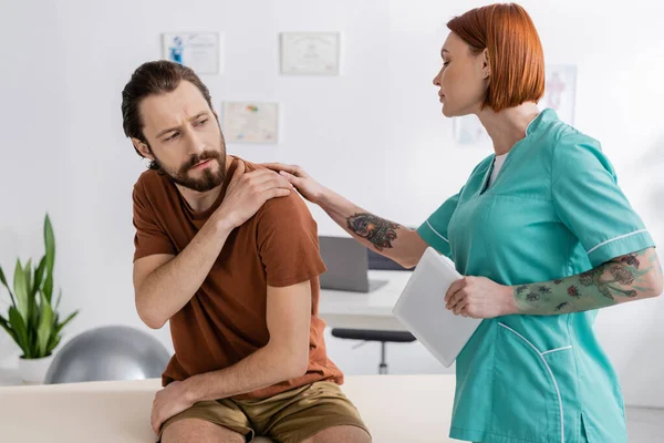 Tattooed physiotherapist with digital tablet touching injured shoulder of bearded man during appointment in rehab center — Stock Photo