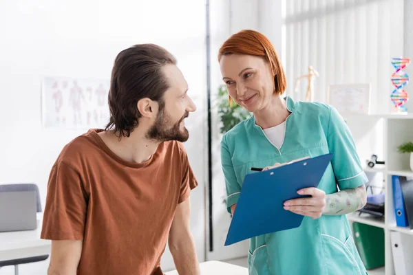 Smiling physiotherapist with clipboard talking to bearded man during consultation in rehabilitation center — Stock Photo