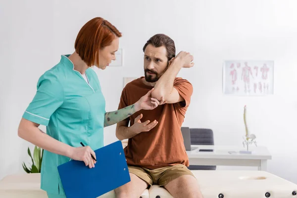 Tattooed physiotherapist with clipboard examining injured elbow of bearded man sitting on massage table in rehab clinic — Stock Photo