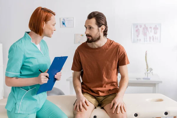 Redhead rehabilitation therapist with clipboard talking to bearded man during appointment in consulting room — Stock Photo