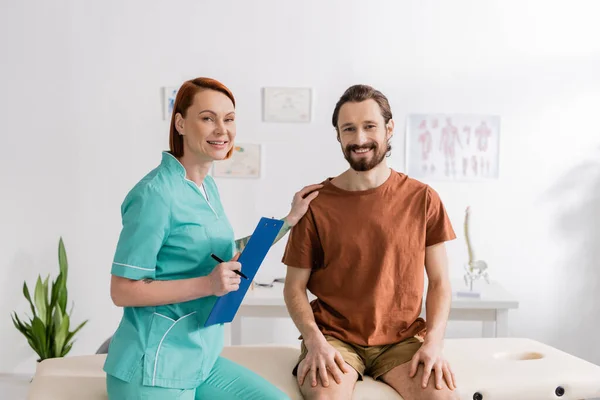 Homme barbu gai et rousse physiothérapeute avec presse-papiers souriant à la caméra dans la salle de consultation — Photo de stock