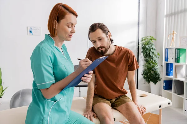 Redhead physiotherapist writing prescription on clipboard near bearded patient sitting on massage table in consulting room — Stock Photo