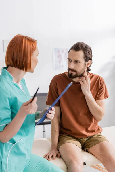 Physiothérapeute rousse avec presse-papiers parlant à un homme barbu touchant le cou blessé dans une salle de consultation — Photo de stock
