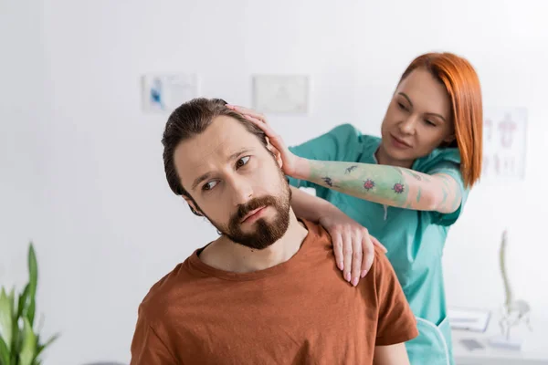 Татуйований фізіотерапевт розтягує шию бородатого чоловіка під час діагностики в консалтинговій кімнаті — стокове фото