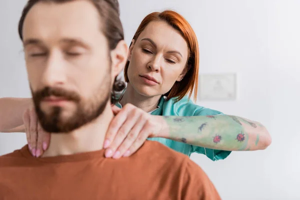 Татуированный физиотерапевт осматривает травмированную шею размытого бородатого мужчины в консультационной. — стоковое фото