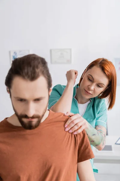 Физиотерапевт, осматривающий травмированное плечо бородатого мужчины в реабилитационном центре — стоковое фото