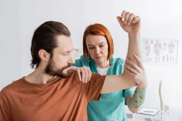 Pelirroja fisioterapeuta flexión brazo y tocando hombro de hombre lesionado en la sala de consulta - foto de stock