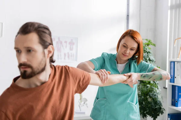 Fisioterapeuta pelirroja estirando el brazo del hombre borroso durante el diagnóstico en el centro de rehabilitación - foto de stock