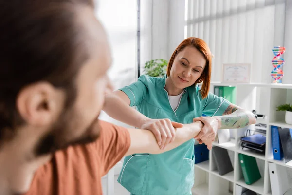 Positive physiotherapist examining injured arm of blurred man in consulting room — Stock Photo