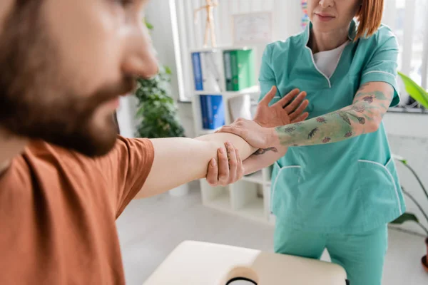 Частичный вид татуированного физиотерапевта растягивающего руку бородатого человека на размытом переднем плане в консультационной комнате — стоковое фото