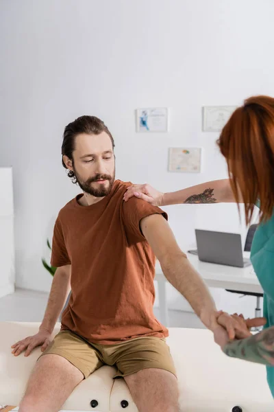 Fisioterapeuta pelirroja examinando brazo de hombre barbudo herido en clínica de rehabilitación - foto de stock