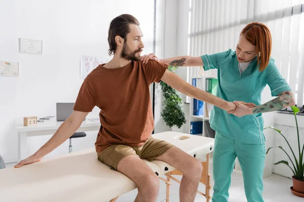 Fisioterapeuta pelirroja examinando brazo lesionado del hombre barbudo sentado en la mesa de masaje en la sala de consulta - foto de stock